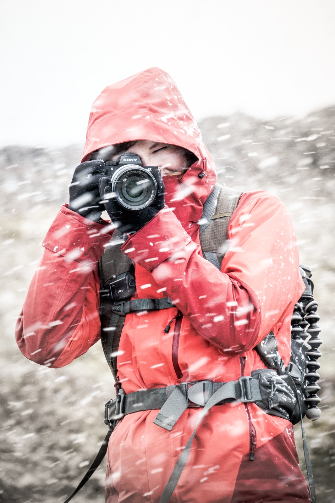 A photographer (Enna Bartlett) in a red coat stands with a camera to her eye while taking a picture in a snowstorm.